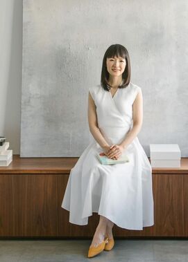 Japanese organizing + decluttering guru Marie Kondo, wearing a white linen dress and seated on a built-in contemporary bench in a minimalist room with gray wall behind her.